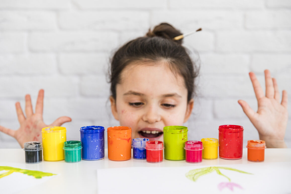 girl-standing-behind-the-table-looking-at-colorful-paint-bottles